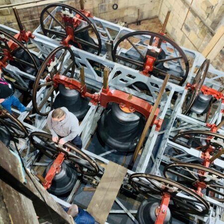 birds eye view inside a belfry, there are 12 bells with red headstocks in a grey frame. there are people standing at the side, listening to a guide who is standing on the bell frame