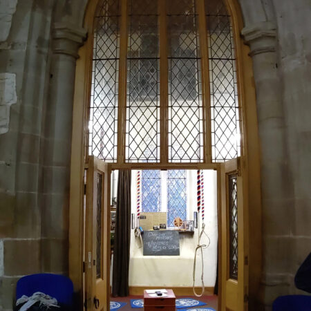 Gothic archway filled with wooden frame screen with diamond pane glass. The door is open, looking into the ringing room. Blue and white mats on the floor, wooden boxes in centre, bell ropes hanging.
